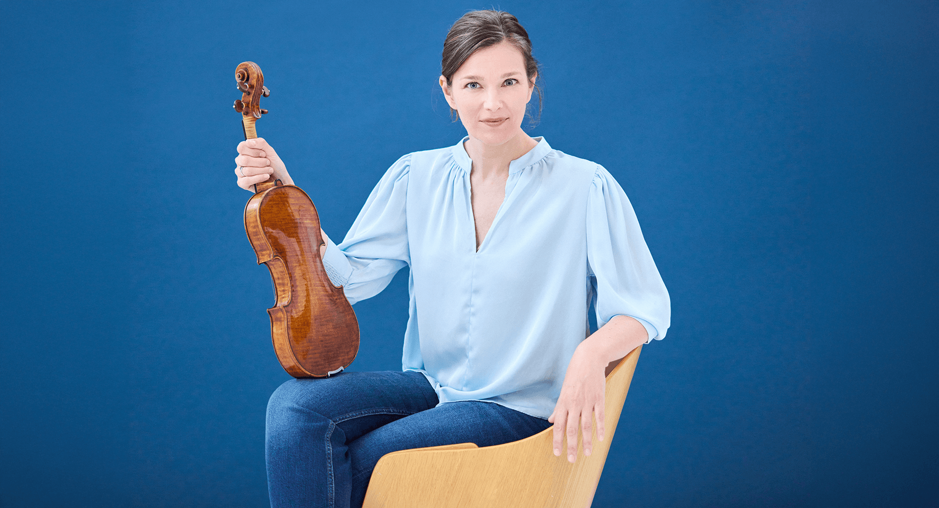 Janine Jansen sitter i en stol med benen i kors och lutar sin fiol mot sitt knä.